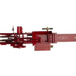 Red metal hay press model with turning gears and springs and four yellow wheels. Top view.