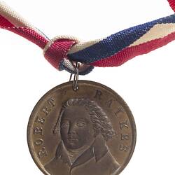Round copper medal with with three-quarter bust of male facing left. Text around. Suspended from ribbon.