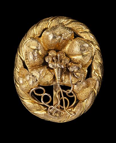 Oval decorative gold brooch containing a flower inside a larger flower, all within an oval border entwined with gold ribbon.
