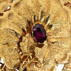 Detail of gold oval locket brooch. Vine leaves and tendrils border. Branches at base with a small bird at top, all supporting a large flower with an amethyst purple paste stone centre.