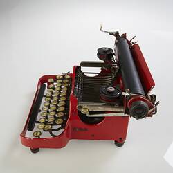 Red metal typewriter, 32 keys, black plastic spacebar at front, black roller for paper at top back. Side view.