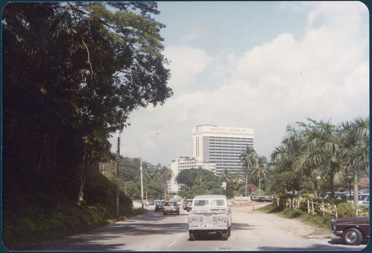 Tall hotel in the distance along a major street, trees line left side and palm tress line right side.