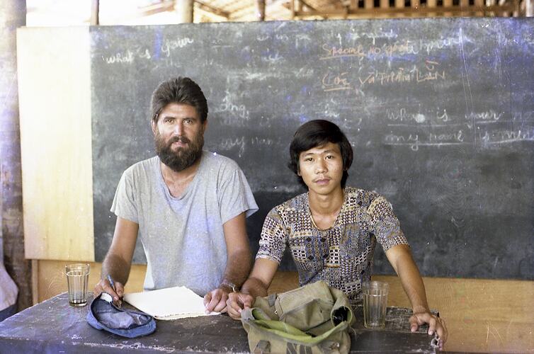 Teo seated men pose. Black chalkboard behind them.