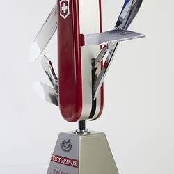 Model of muti-tool on stand. Red handle with all tools out. Large blade, short blade, scissors, bottle opener (with flathead screwdriver), a can opener (with flathead screwdriver), and a reamer.