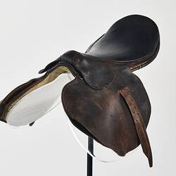 Horse racing saddle. Padded dark brown leather. Double layer beneath saddle flap. Two leather straps on either side. Three quarter front view.
