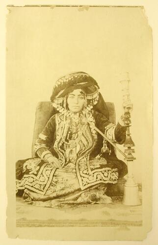 Seated Kurdish woman in traditional clothing, holding a kalyan.