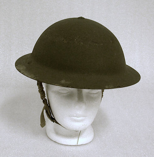 Painted khaki coloured metal helmet with chin strap, on white mannequin head.