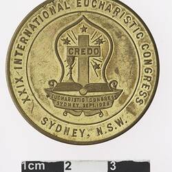 Round gold coloured medal with cross and chalice, above book and banner, text surrounding.