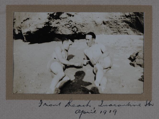 Two men in bathing trunks reclining on sand, with rocks behind.