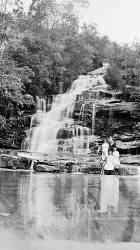 Group of people sitting by a lake at the base of a waterfall.