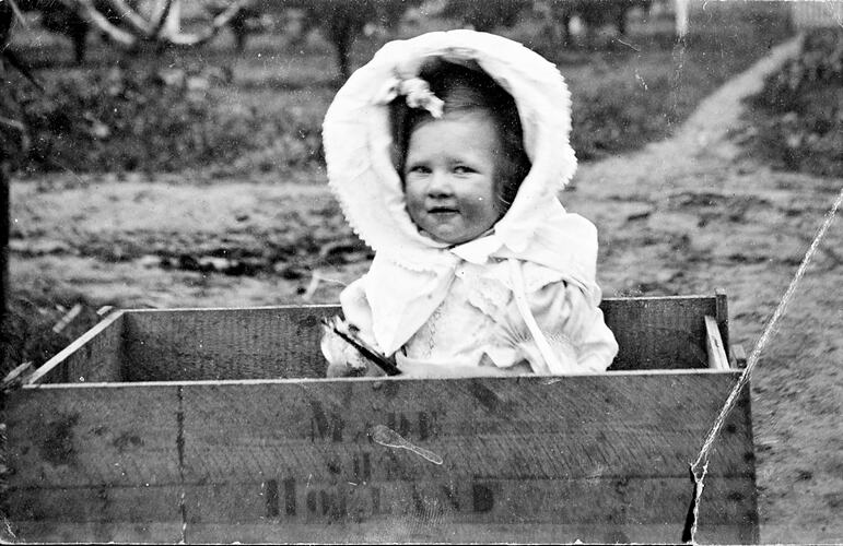 Young girl wearing white bonnet sitting in wooden fruit crate.