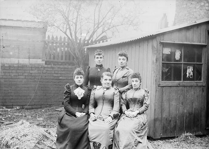 Five women in Victorian dress posed in a backyard near a garden shed.