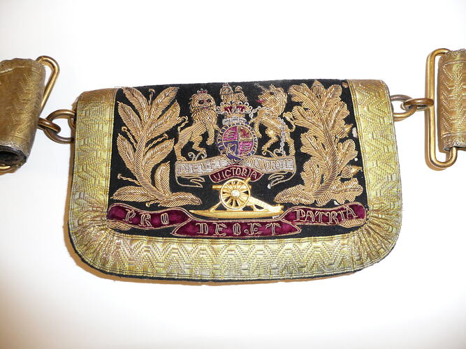 Black pouch attached to belt, fabric flap embroidered in gold with coat of arms above cannon and red banner.