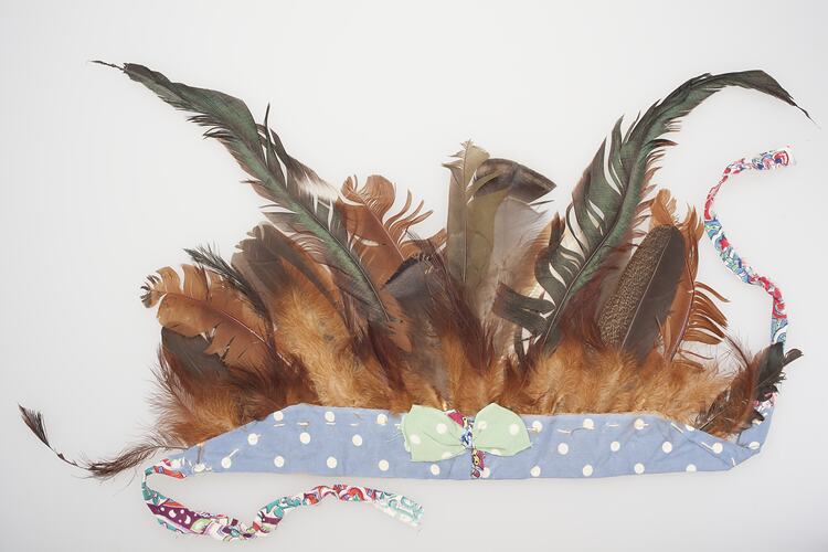 Feathered headdress. Brown feathers mounted in a blue and white spotted band with green bow.