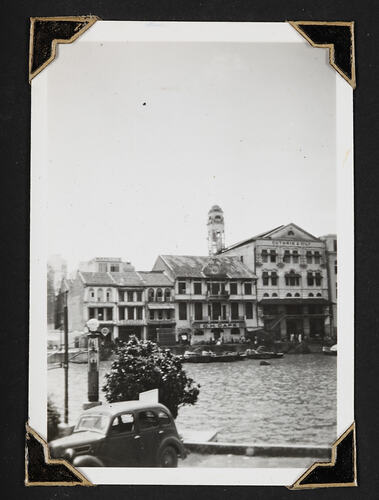 Buildings besides river with small boats in front and car in foreground.