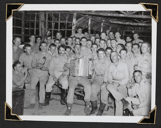 Group of seated men with man in centre playing accordion.