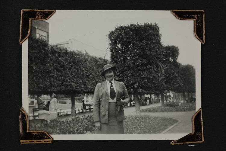 Woman standing on a street, tree hedges and people sitting on park benches behind.