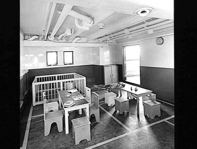 Ship interior. Children's playroom. Two small tables and chairs at front. Playpen behind at left.