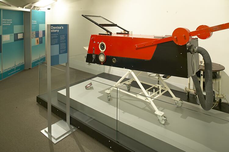 Red rectangular metal box with opening at one end and tube fitted to other end. On white wheeled stand.