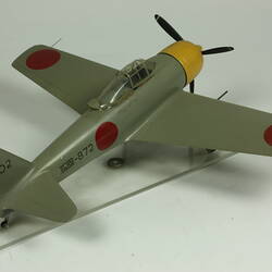 Green grey plane model with red spot on each wing and fusleage. Yellow nose, black propeller. Back view.