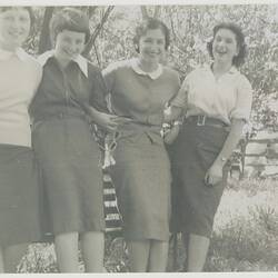 Four smiling women wearing pencil skirts with blouses, jackets or cardigans  lean against a park bench seat.