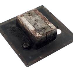 Black metal square (burnt) with holes around edges and two larger ones within. Grey metal rectangular in centre.