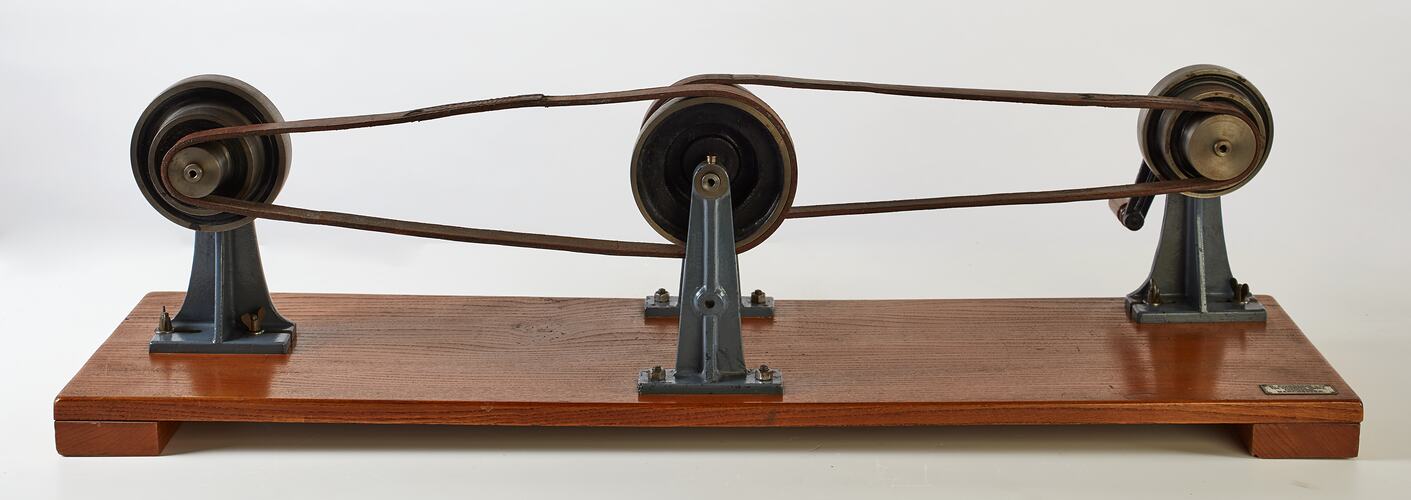 Wooden base with two pairs of round wheels, each connected via a pulley looped belt. One wheel has a handle.