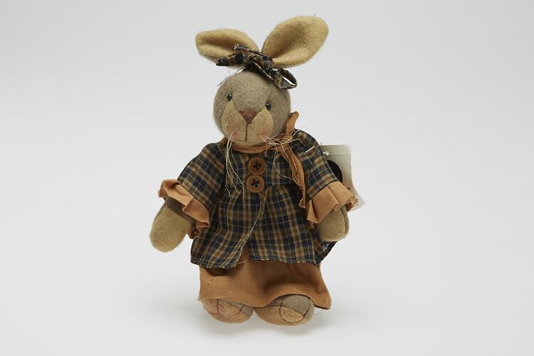 Soft toy rabbit dressed in brown checked dress. Has long whiskers.