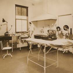 Medical room with examination table in centre, nurse standing at sink and cupboard filled with medicines.