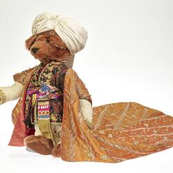 Light brown plush bear wearing white turban, ornate tunic and coat with gold embroidery. Coat has long tail.