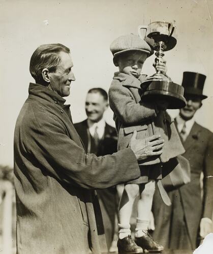 Man supports a young boy holding up a horse racing trophy. Onlookers smile.