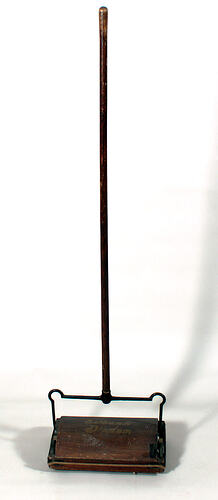 Carpet sweeper, wooden handle and sweeper pan, shown upright. Base has gold text.