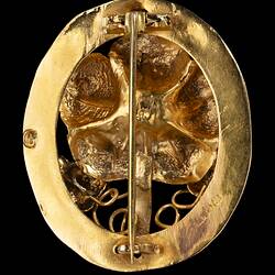 Back of oval decorative gold brooch containing a flower inside a larger flower, all within an oval border entwined with gold ribbon.