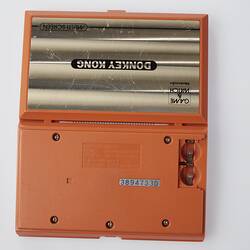 Open facedown orange plastic, handheld game console. Top (front) has metallic label/text. Battery spot in base