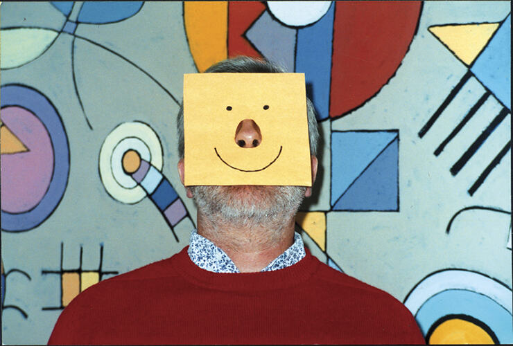 Man with a yellow square piece of paper on his face with a cut-out section for his nose. It has eyes and a mou