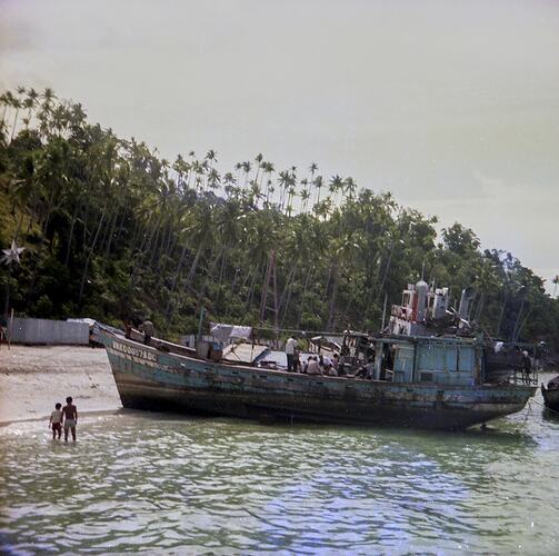 Crowded refugee boat moored at the beach. Heavily treed hill in background.
