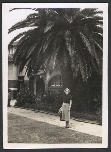 Woman standing on pathway posing in front of a building with an arched window and palm tree. Grass in foreground.
