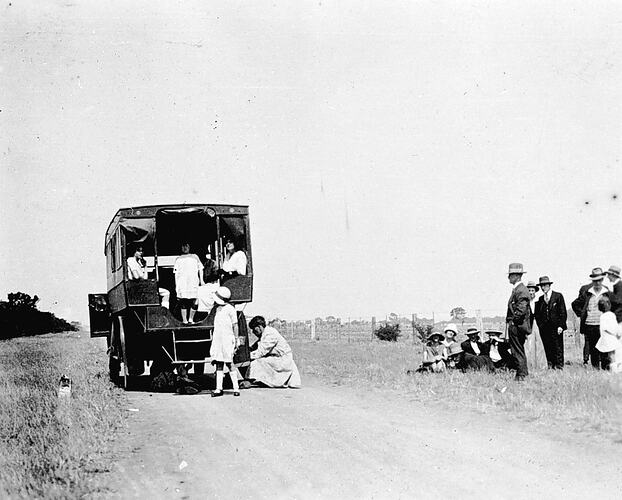 [A passenger charabanc halted by a flat tyre, Ballarat, about 1920. Most of the passengers wait beside the road while some mend the tyre.]