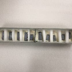 Long narrow box of 10 rectangular metal weights. Each is pierced at top.