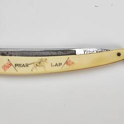 Cut throat razor, metal blade, ivory-styled plastic handle with 'PHAR LAP', picture of jockey on racehorse and two flags. Closed.