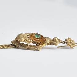 Back of gold pendant earring. Comprised of four connected sections with floral and leaf motifs. Gold fringe hangs from the bottom. Profile.