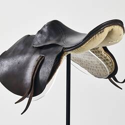 Horse racing saddle. Padded dark brown leather. Double layer beneath saddle flap. Two leather straps on either side. Three quarter front view.
