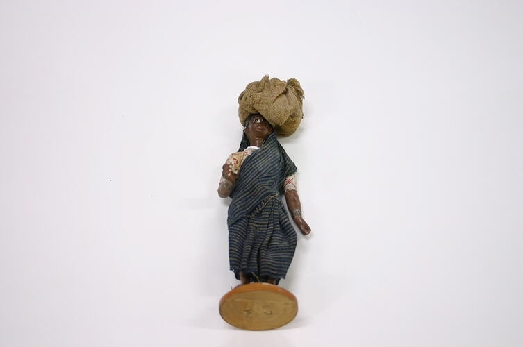 Figurine of an Indian man in traditional dress carrying a large sack on his head.