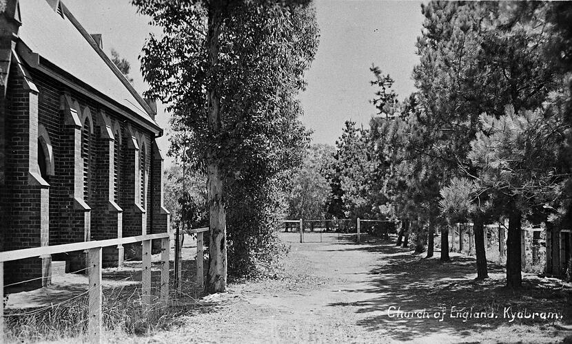 Tree lined dirt driveway with part of brick wall of church visible on left.
