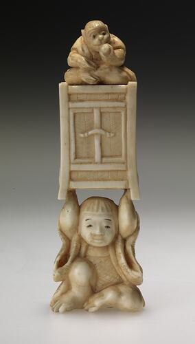 Seated boy lifting a box above his head. Atop the box is a monkey eating fruit.