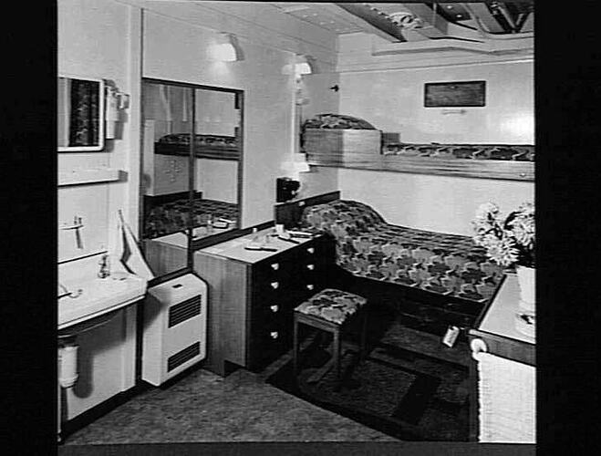 Ship interior. Single bunk beds at back against wall. At left handbasin and chest of drawers at right.