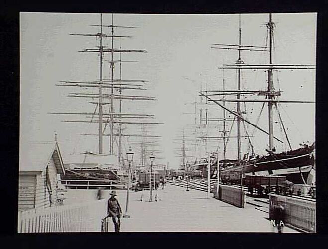 Freight being unloaded from a number of sailing ships moored at a pier.