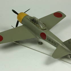 Green grey plane model with red spot on each wing and fusleage. Yellow nose, black propeller. Back view.