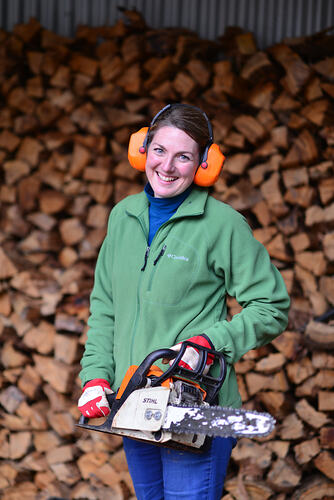 Woman standing near stack of wood and holding a chainsaw.