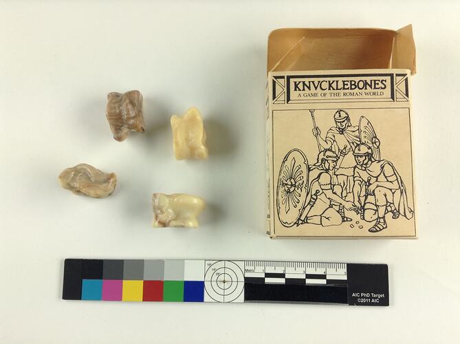 Four cream plastic numbered knucklebones in cardboard box with illustration of Roman soldiers.